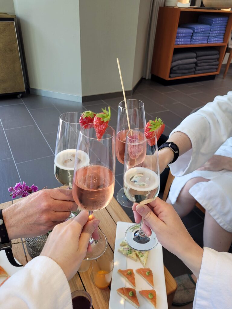 Vier Champagner-Gläser zusammen gehalten, dekoriert mit Erdbeeren, es sind nur die Hände zu sehen inkl. ein wenig des Bademantels