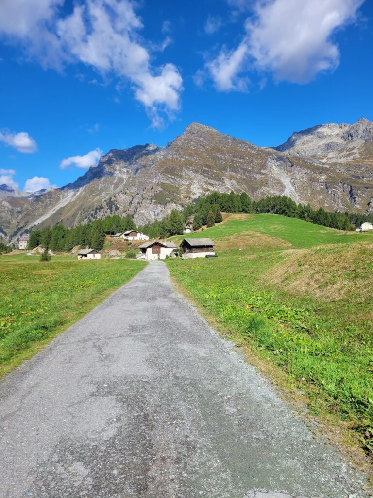 Engadin Maloja Strasse, die in einen kleinen Weiler führt, im Hintergrund Berge und blauer Himmel
